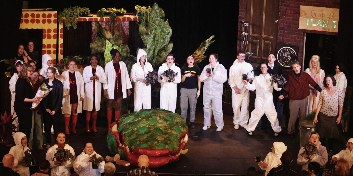 Dr. Kimberly Francis, professor and director, School of Fine Art and Music, College of Arts, University of Guelph, stands on stage at War Memorial Hall, holding a bouquet. Beside her is the "Little Shop of Horrors" student ensemble standing around Audrey II, the symbolic carnivorous plant from the production.